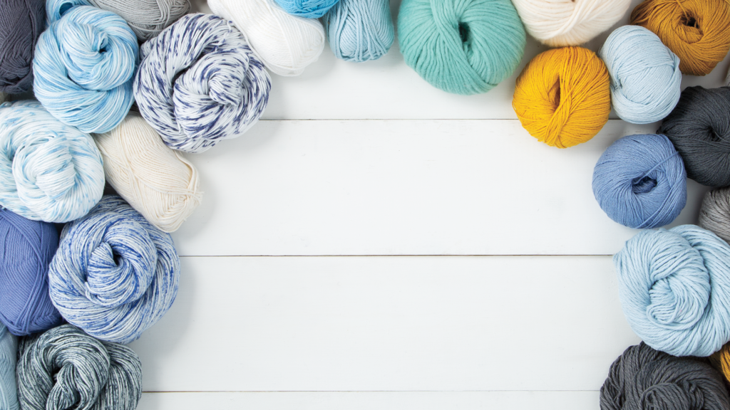 A white wooden plank background with an arc of yarn across the sides and top, in blue, white, green, yellow, and gray tones