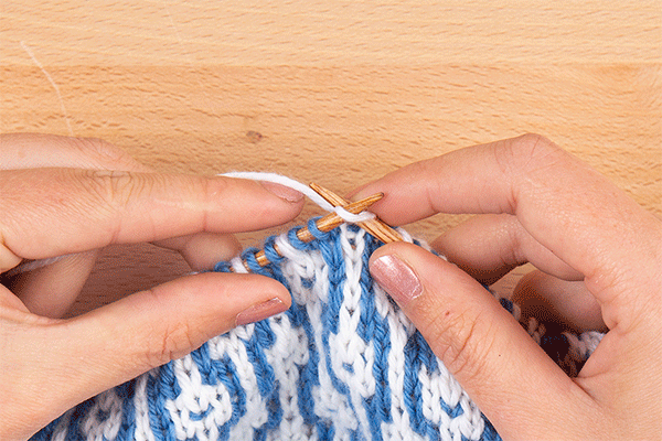animation of four stitches being worked: knit 2, slip 1, knit 1. Image description: a close-up view of hands knitting a mosaic swatch made of medium blue and white stitches.