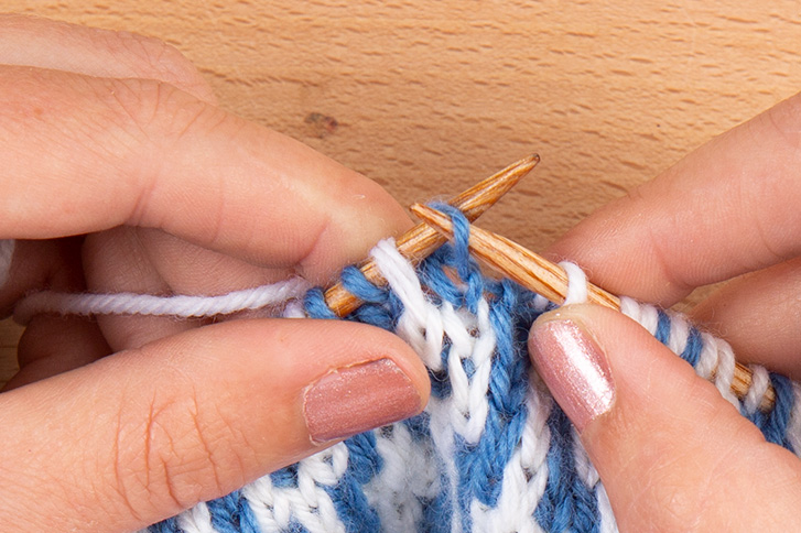 right-hand needle is inserted into stitch on left-hand needle, purl-wise. Image description: a close-up view of hands knitting a mosaic swatch made of medium blue and white stitches.