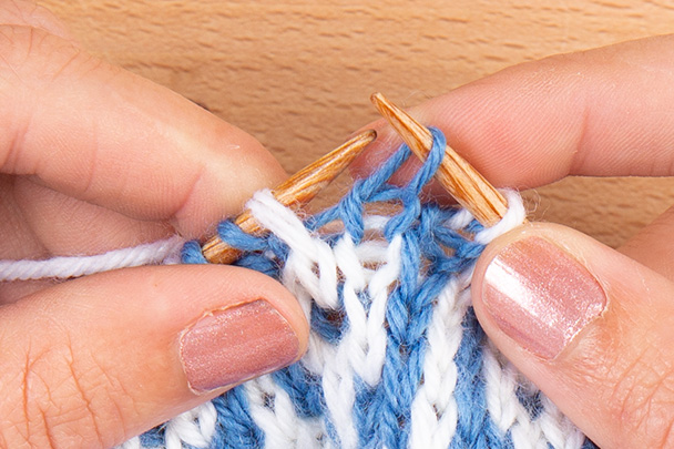 stitch has just been slipped from left-hand needle to right-hand needle. Image description: a close-up view of hands knitting a mosaic swatch made of medium blue and white stitches.