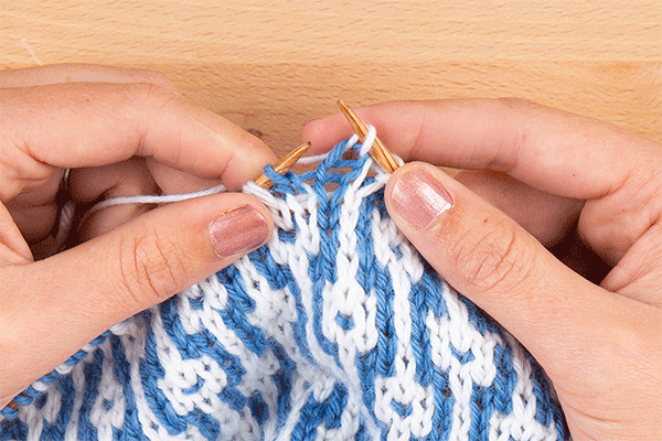 animation of stitch being slipped. Image description: a close-up view of hands knitting a mosaic swatch made of medium blue and white stitches.