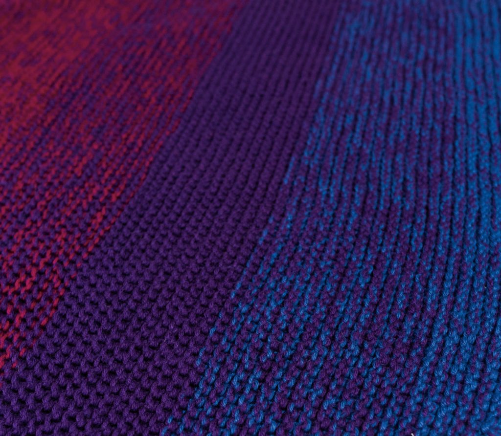 Knit Picks 12 Weeks of Gifting Patterns, featuring the Continuation Blanket. Image shows: a closeup of the knitted texture of the blanket. The blanket gradates from magenta, to purple, to blue, along the diagonal.